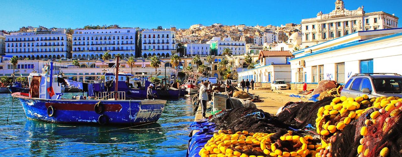 A bright blue fishing boat moored at a dock in Casbah, Algiers, surrounded by nets and ropes, with calm Mediterranean water reflecting the boat and nearby harbor structures.