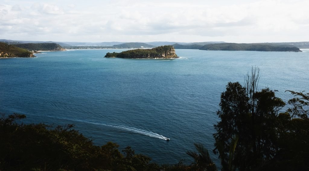 mall rocky island sitting in calm blue water inside Ku‑ring‑gai Chase National Park, surrounded by gentle morning light. The scene feels peaceful and remote, with soft grey skies, still water and rugged bushland framing the bay.