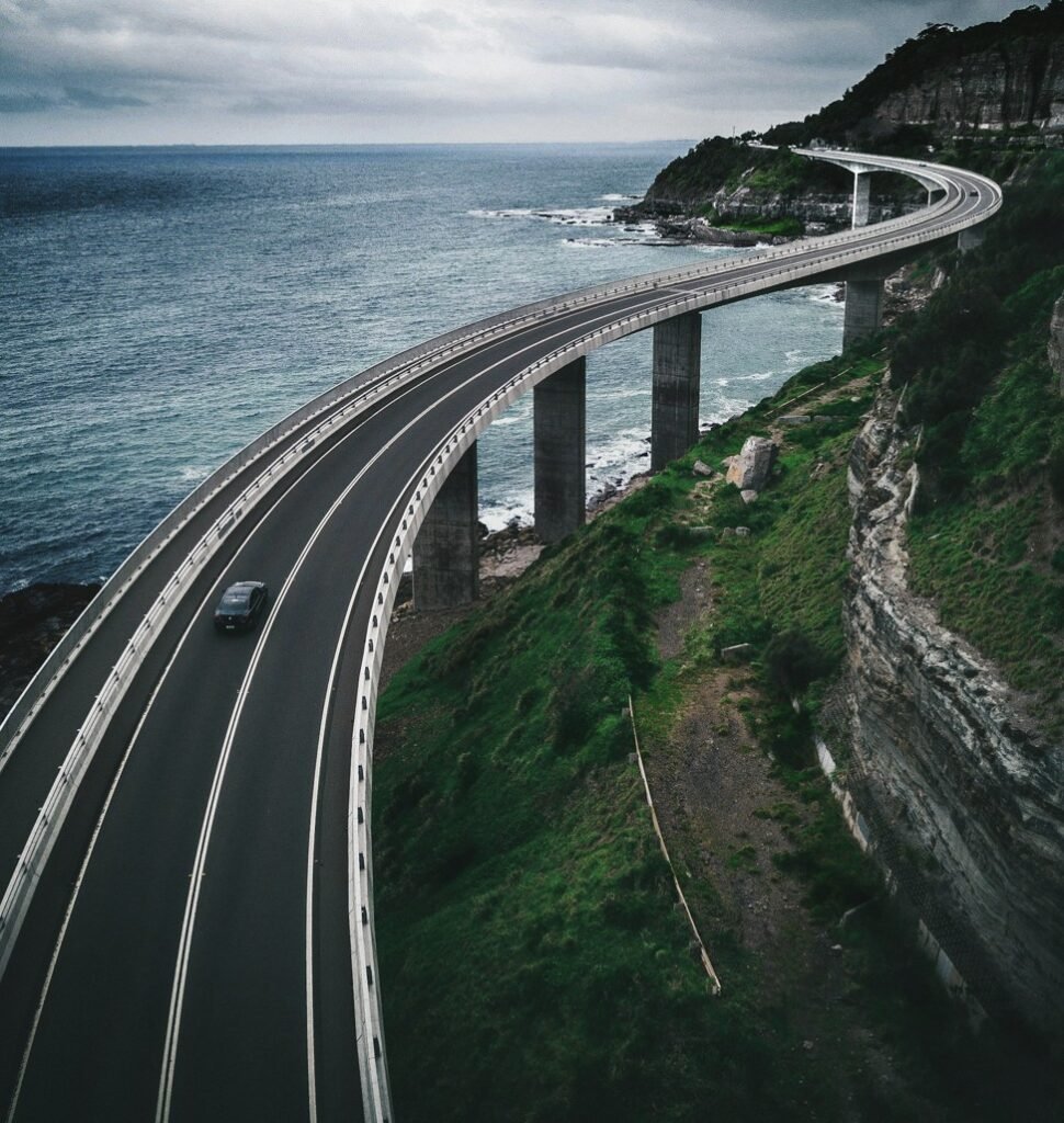 The cliffs rise sharply beside the bridge, covered in patches of grass and rugged rock. Above, the sky is bright with soft white clouds, giving the whole scene a dramatic yet peaceful coastal atmosphere.