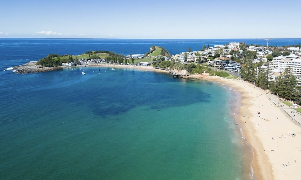 This aerial photo shows Terrigal Beach on the NSW Central Coast, with golden sand curving along the shoreline, turquoise water rolling in gentle waves, and the town’s coastal buildings lining the edge of the beach. The scene captures Terrigal’s classic mix of ocean beauty, relaxed holiday energy and a vibrant seaside strip.