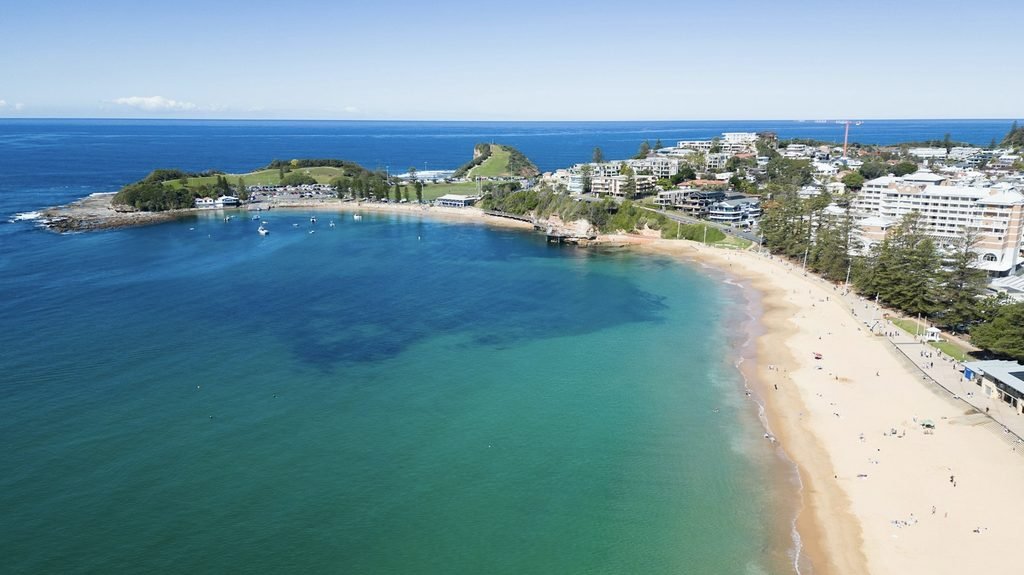 A high-angle aerial photograph of Terrigal Beach in New South Wales, Australia, showing golden sand curving along the coastline, turquoise water rolling onto the shore, and the surrounding coastal town stretching inland.