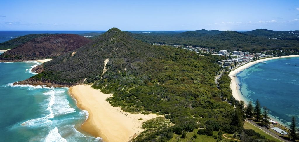 This aerial photo shows Tomaree National Park in Shoal Bay, Port Stephens (NSW). You can see bright turquoise water meeting a long curve of sandy shoreline, with rugged green headlands framing the bay.