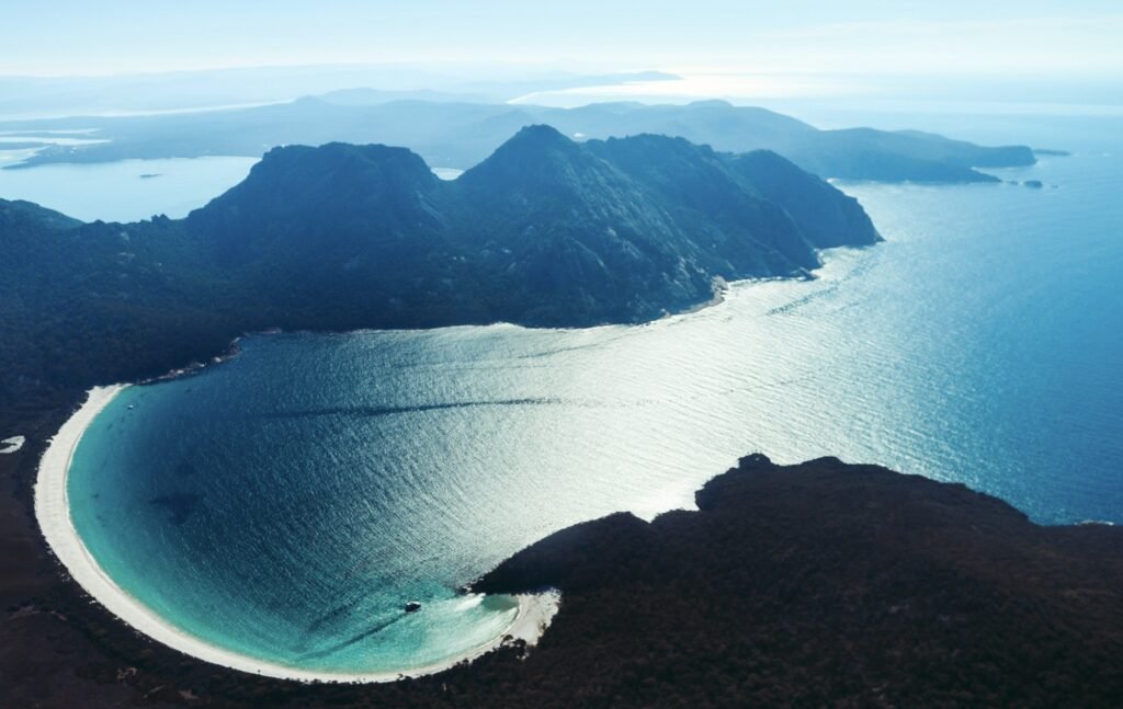 Aerial view of Wineglass Bay in Tasmania, Australia, showing a curved white-sand beach meeting vivid turquoise water, surrounded by rugged forested mountains under clear daylight.