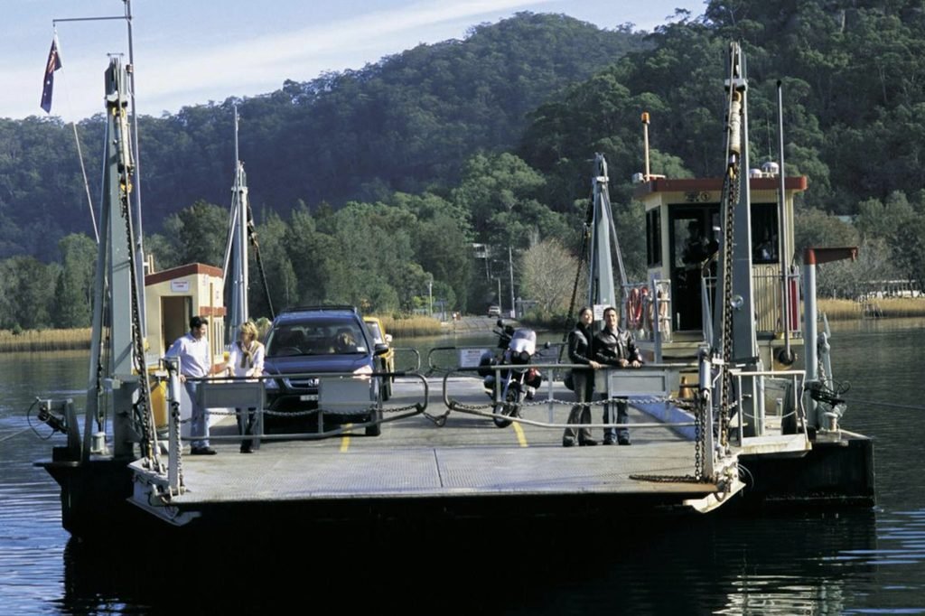 A cable ferry carrying cars and passengers across the Hawkesbury River at Wisemans Ferry, surrounded by forested hills and calm water under a clear blue sky.